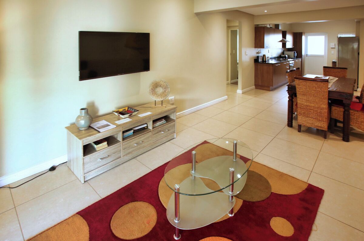 A modern living room with a flat-screen TV, a glass coffee table, and a dining area with wicker chairs.