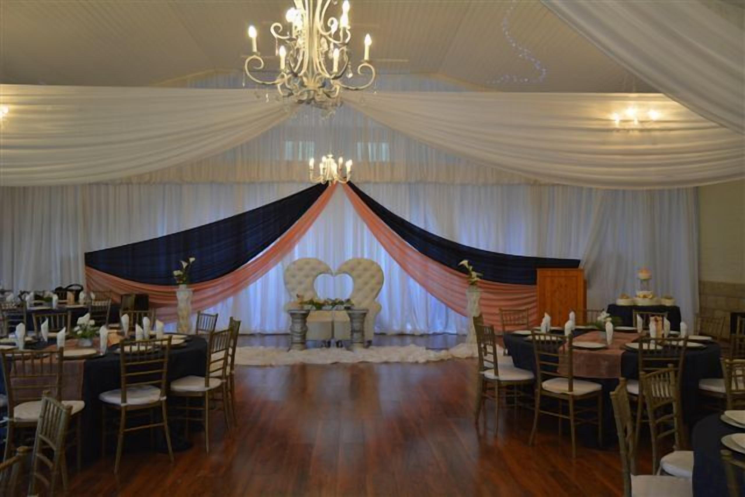 Un salón de banquetes con mesas y sillas redondas, un escenario con tela decorativa y una araña.
