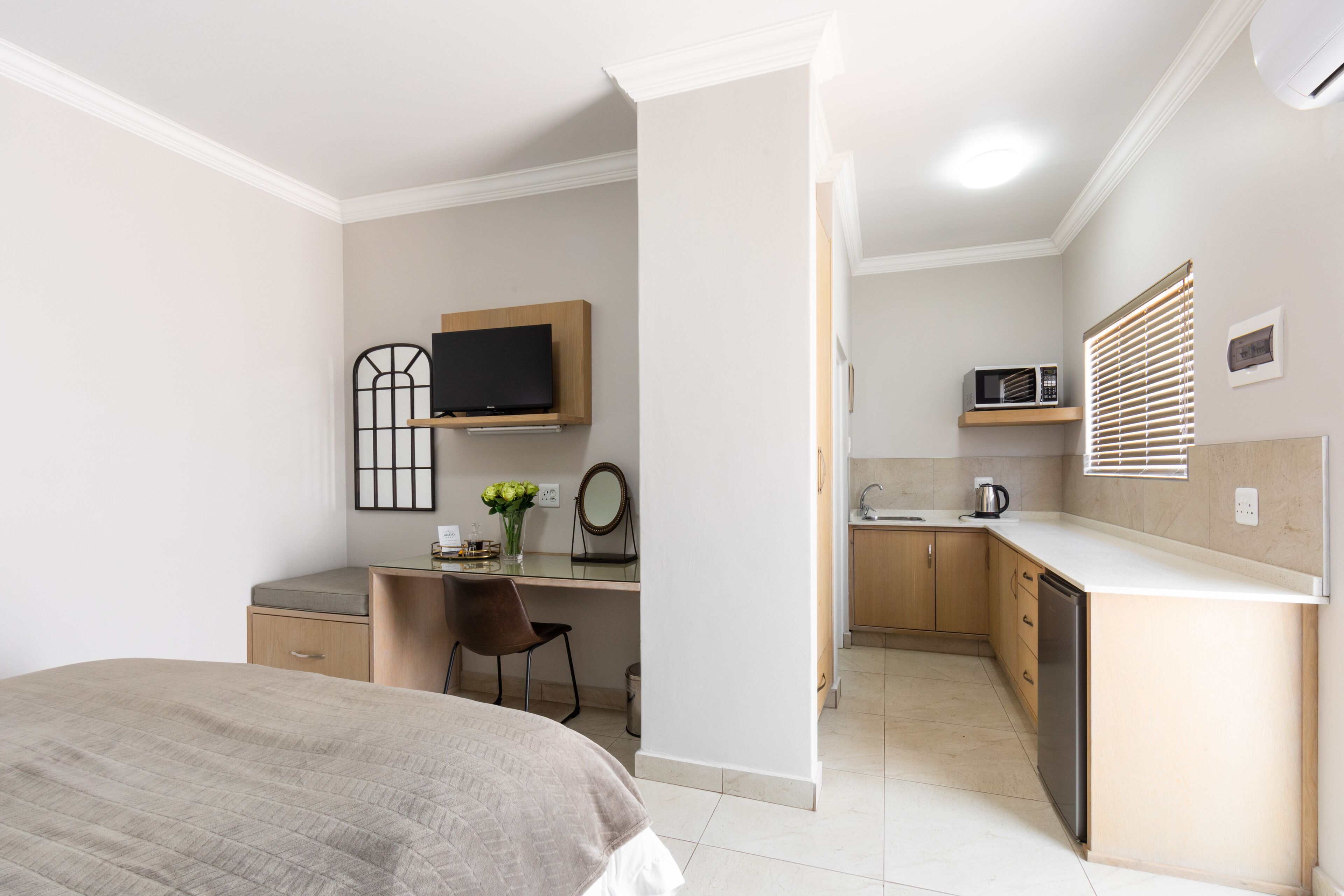 A compact bedroom with a bed, a desk with a chair, a television, and a small kitchenette with a microwave and sink.
