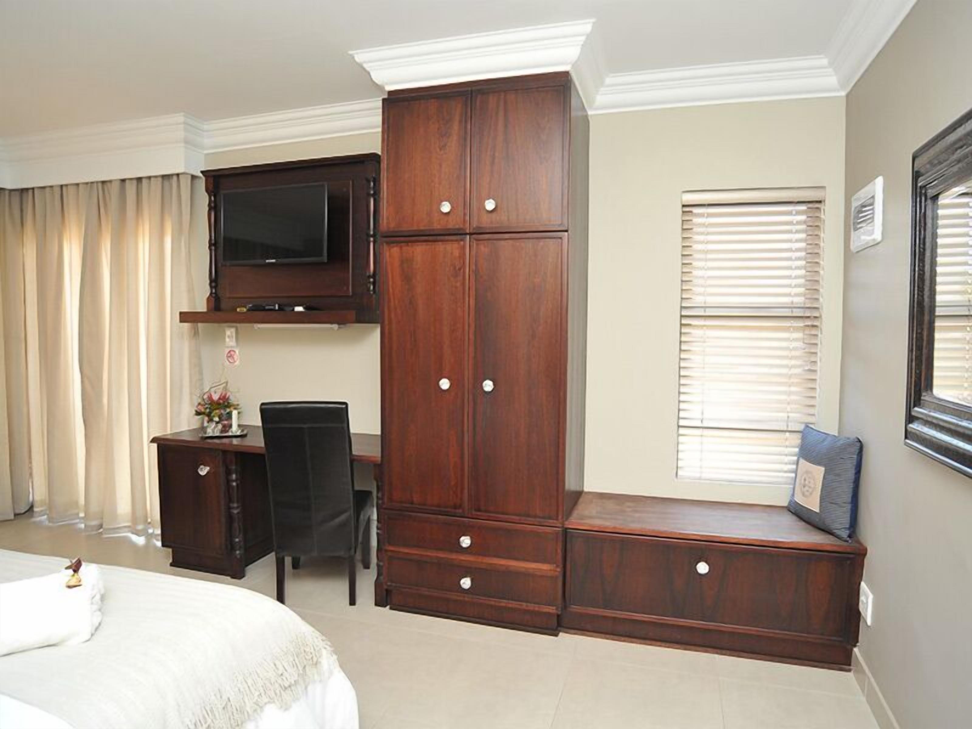 A bedroom with a bed, a desk, a TV mounted above it, a wardrobe, and a window with blinds.