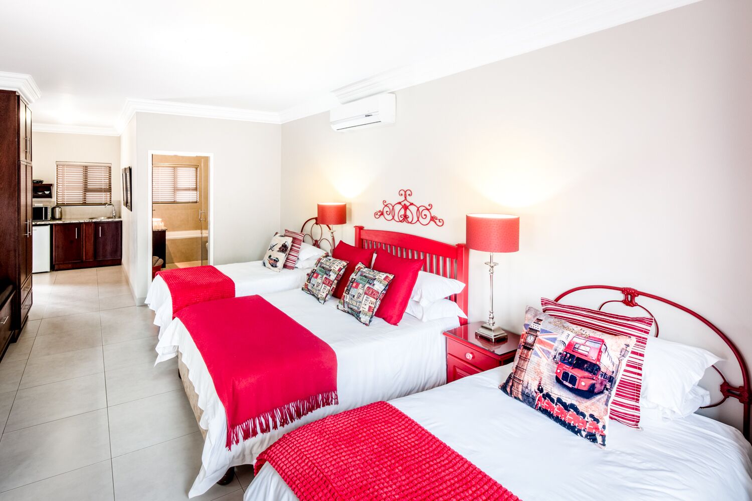 A hotel room with two beds, each with red bedding and white pillows. There is a red headboard, a bedside table with a lamp, and a window with blinds.