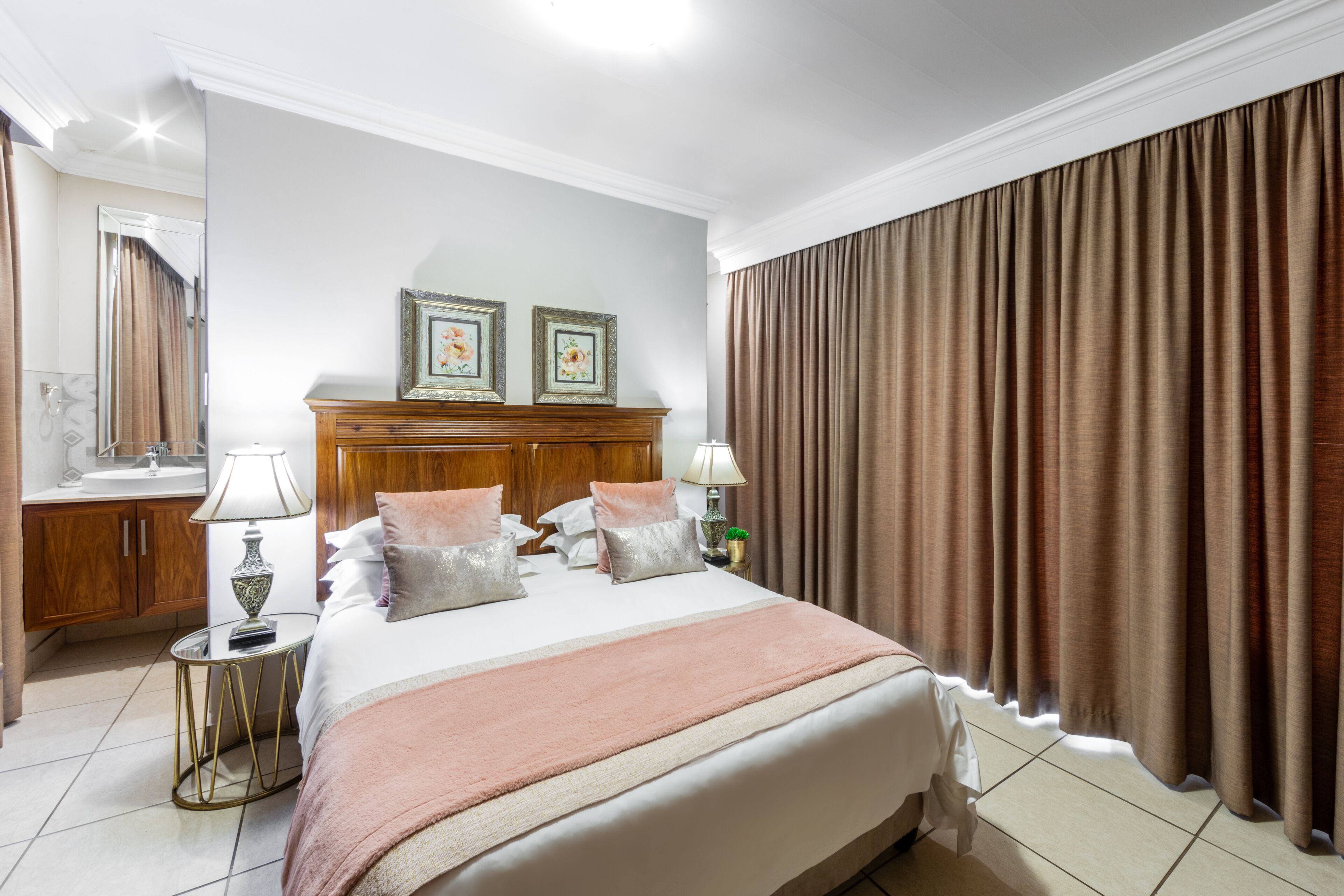 A hotel room with a large bed, a wooden headboard, two bedside lamps, a nightstand, a mirror, and a window with curtains.