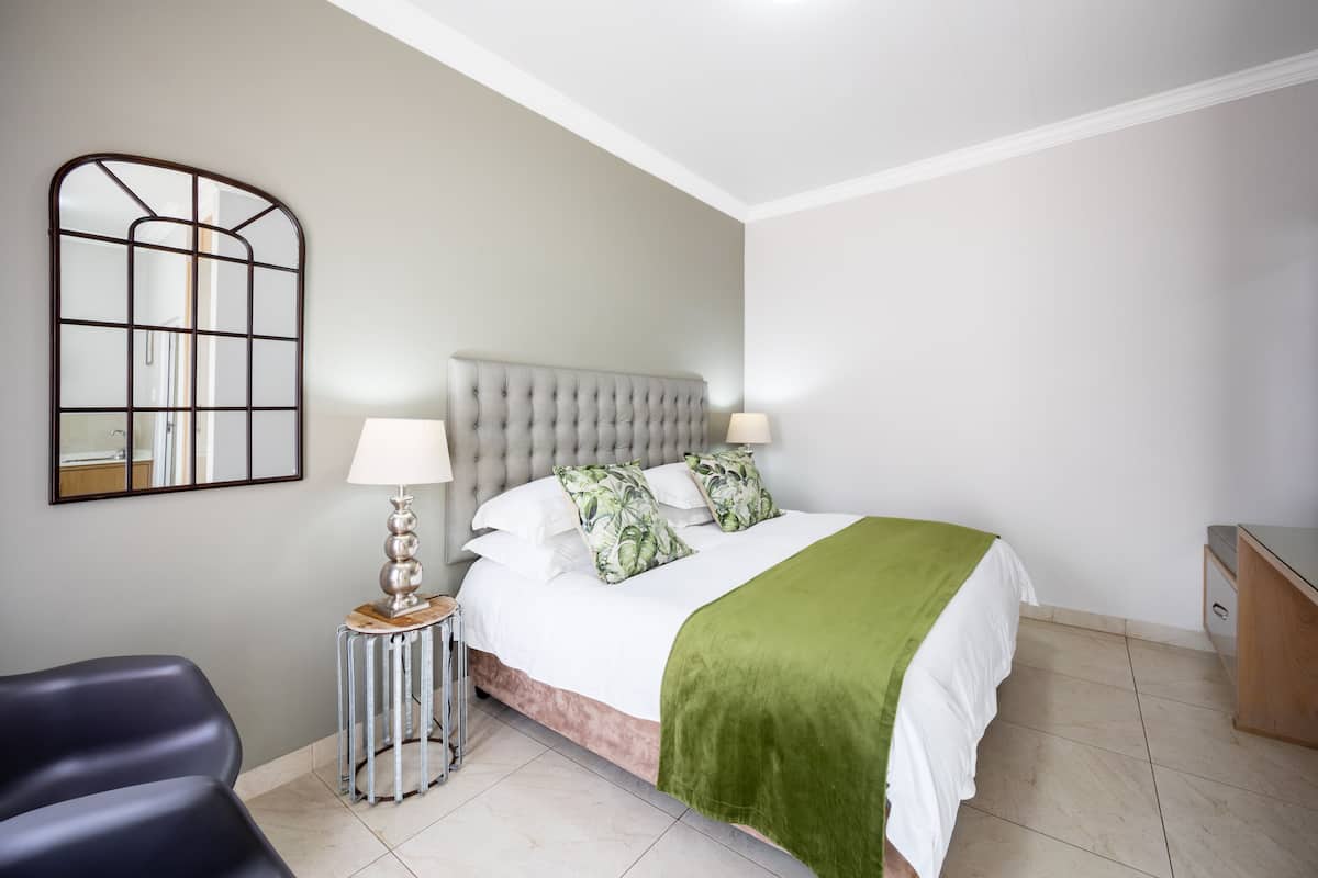 A bedroom with a tufted headboard, a bed with white bedding and a green throw, a bedside table with a lamp, a mirror, and a chair.