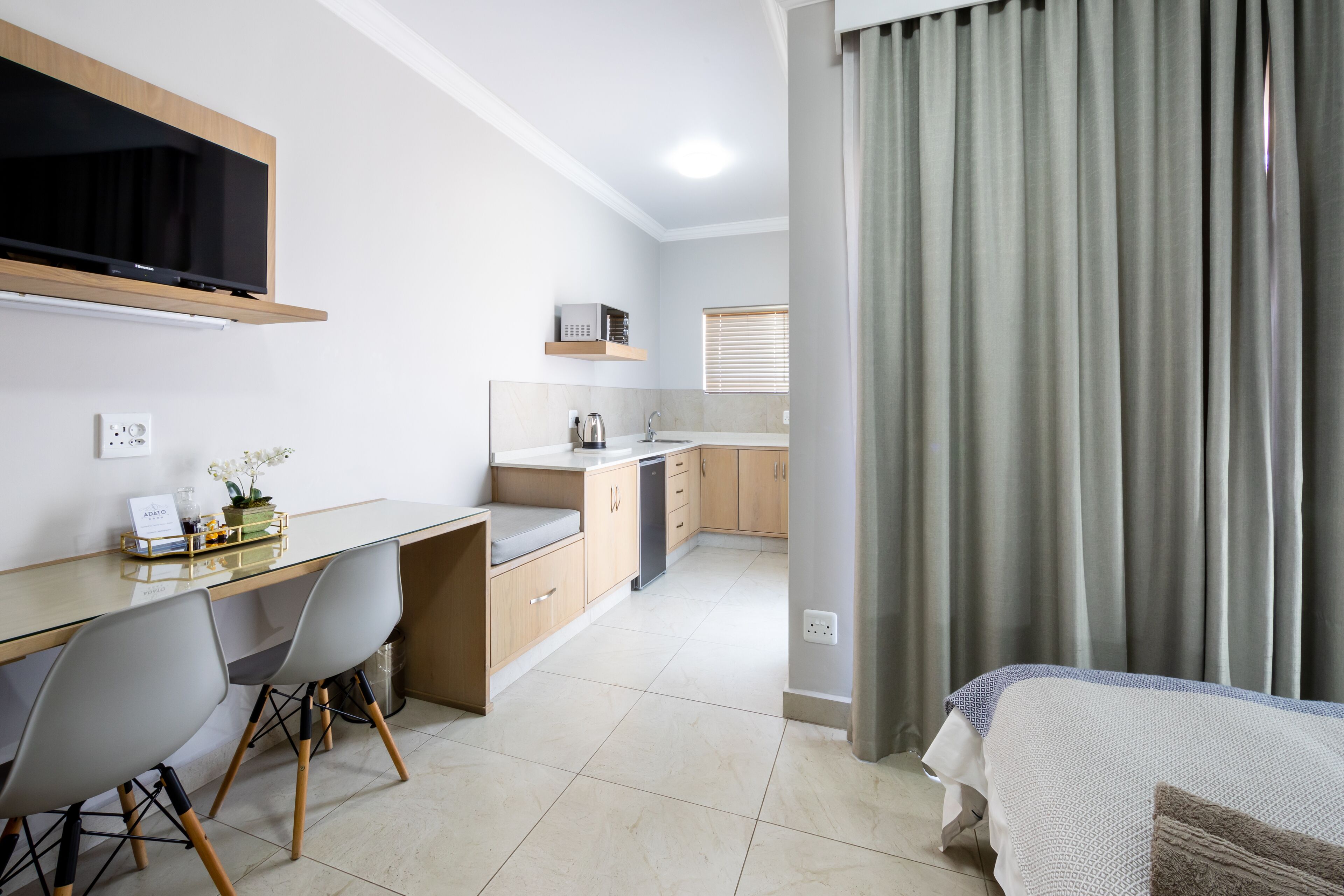 Ein modernes Hotelzimmer mit Küchenzeile, einem Essbereich mit Tisch und Stühlen, einem Bett und einem großen Fenster mit Vorhängen.