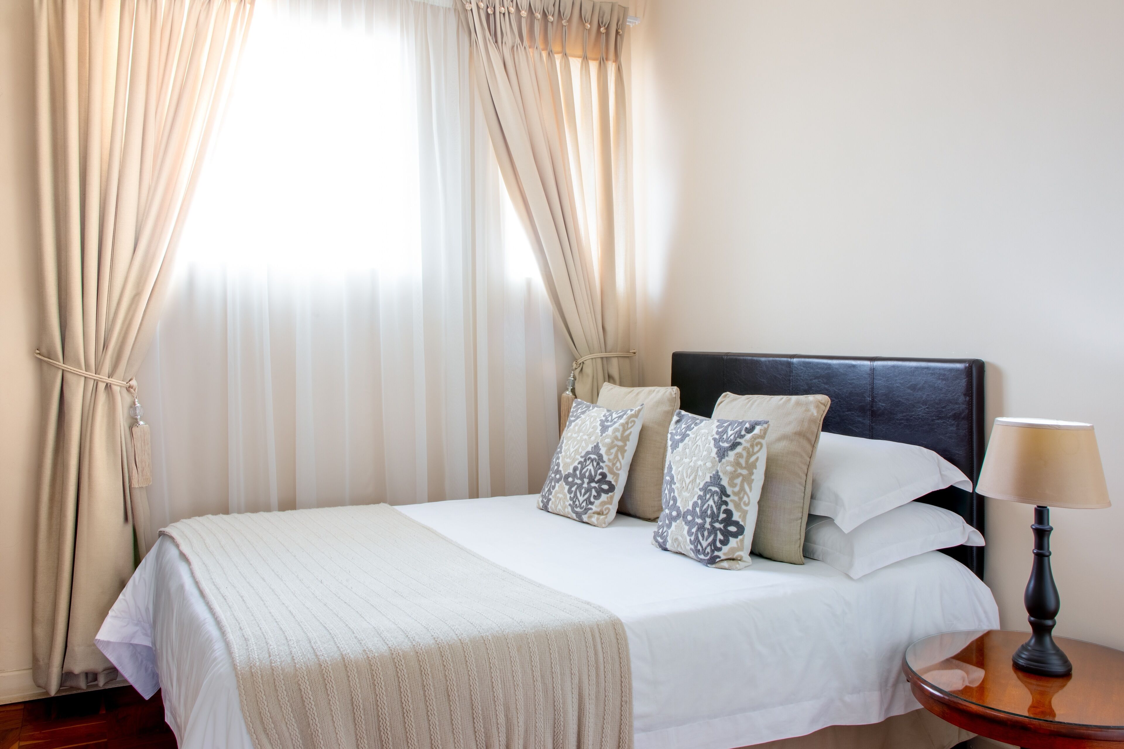 A neatly made bed with a white bedspread and decorative pillows, a bedside table with a lamp, and sheer curtains allowing natural light.