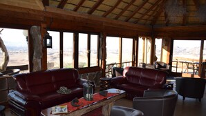 Ein rustikales Interieur mit Holzbalken, Ledersofas, einem Holztisch und großen Fenstern mit Blick auf eine Wüstenlandschaft.