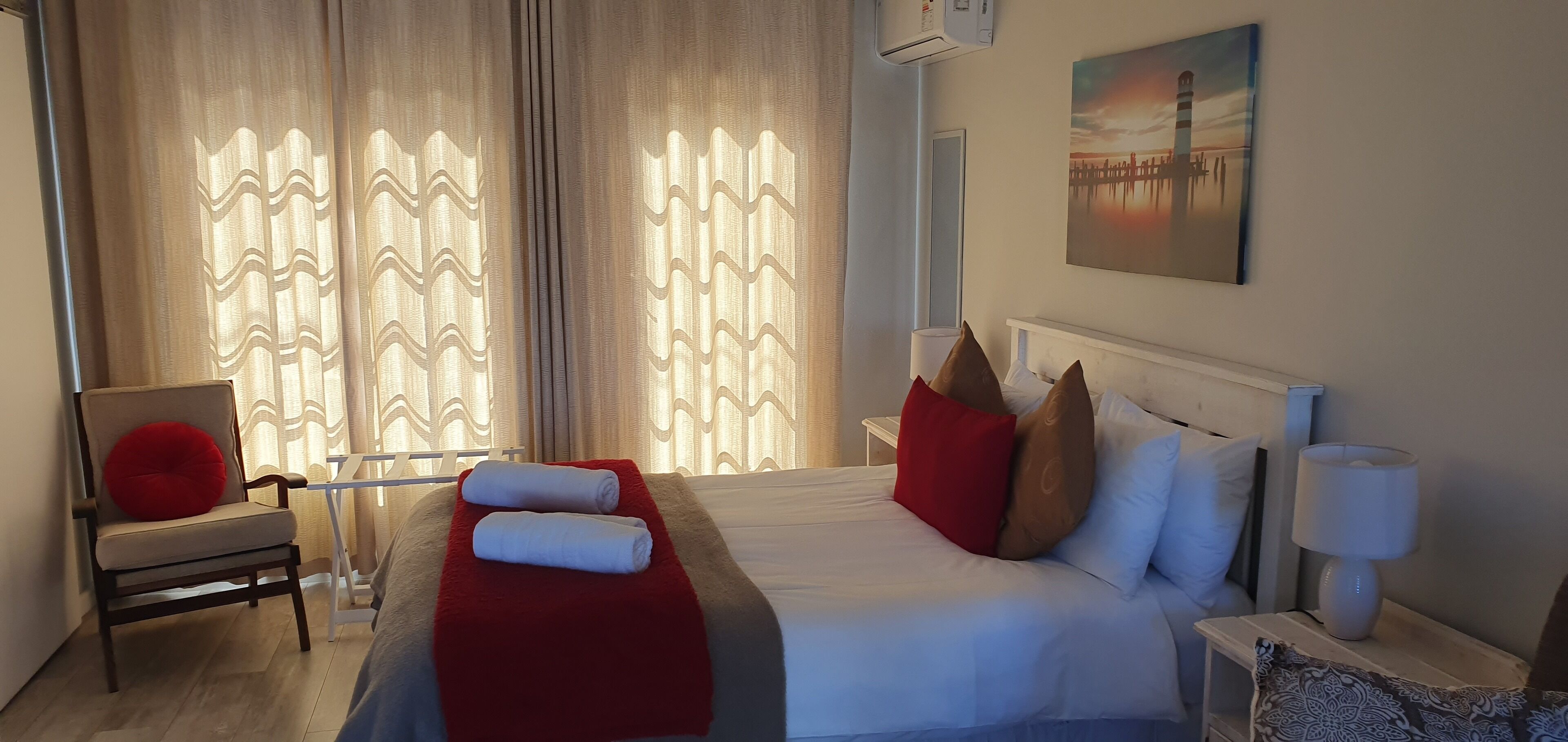 Een hotelkamer met een bed, een stoel, een rood kussen, een witte lamp en een schilderij aan de muur.