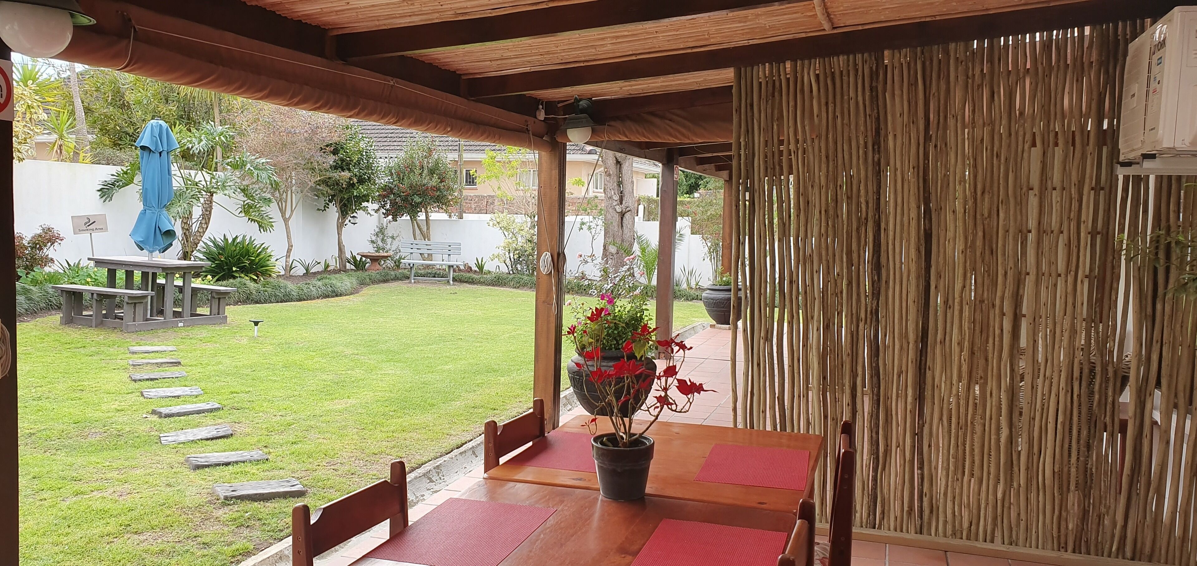 An outdoor patio with a wooden table and chairs, a bamboo screen, and a view of a garden with a bench and a blue sculpture.