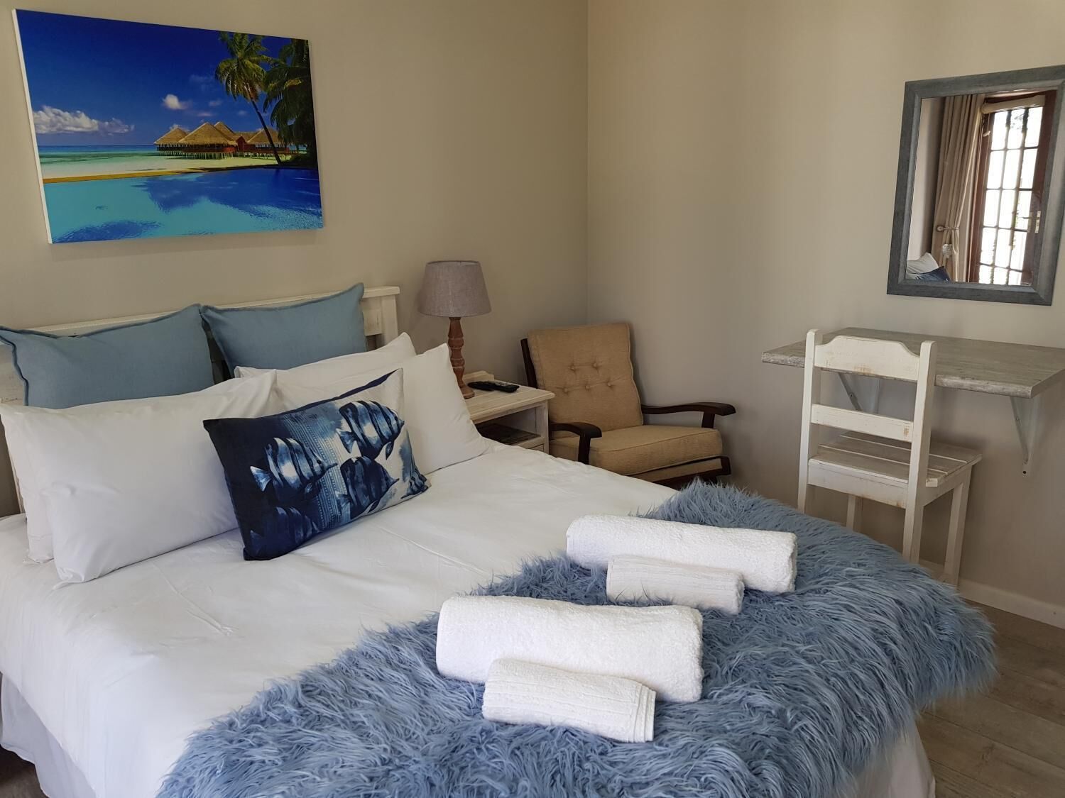 A bedroom with a bed, a chair, a desk, a lamp, and a painting of a beach scene.