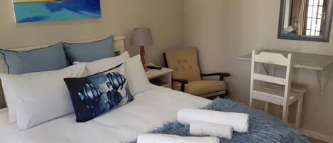 A bedroom with a bed, a chair, a desk, a lamp, and a painting of a beach scene.
