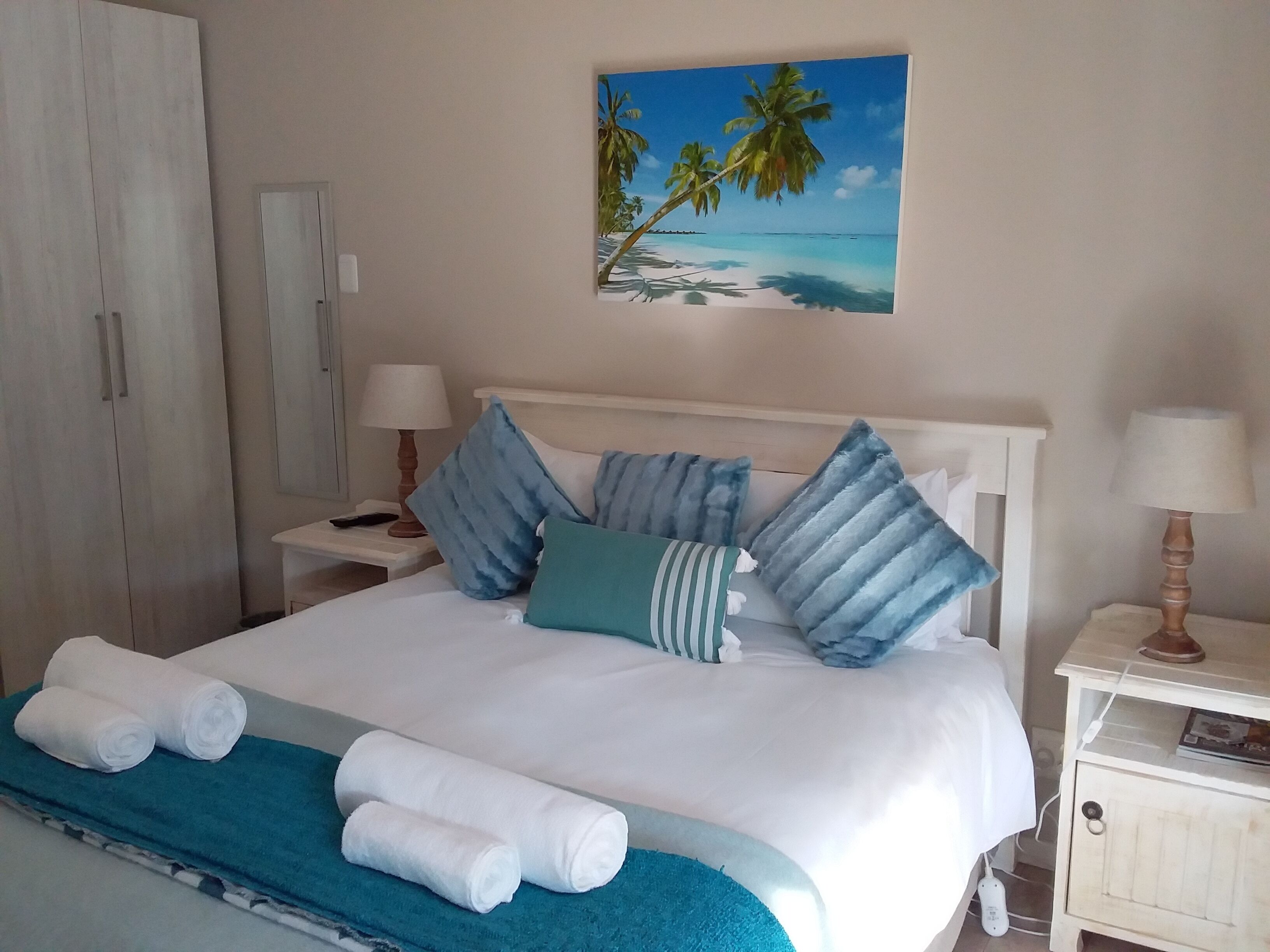 Una cama bien hecha con sábanas blancas y almohadas azul rayadas, una manta azul turquesa y toallas blancas enrolladas sobre un corredor azul para la cama.