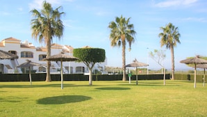 Een gazon met palmbomen en rieten parasols, een wit gebouw met betegeld dak, en een helderblauwe lucht.