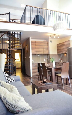 A spacious living area with a staircase leading to a loft, a dining area with a table and chairs, and a kitchen with wooden cabinets. - Apartamenty Baltyckie - Bulwar Portowy (Ustka)