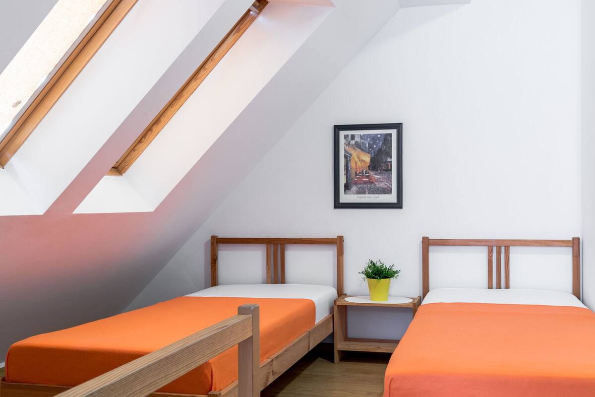 A dormitory room with two wooden bunk beds, a small bedside table with a plant, and a framed picture on the wall.