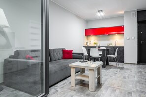 Un salon moderne avec un canapé gris, une table basse en verre et un coussin rouge. On aperçoit à l’arrière-plan un coin cuisine avec des meubles rouges et une porte noire.