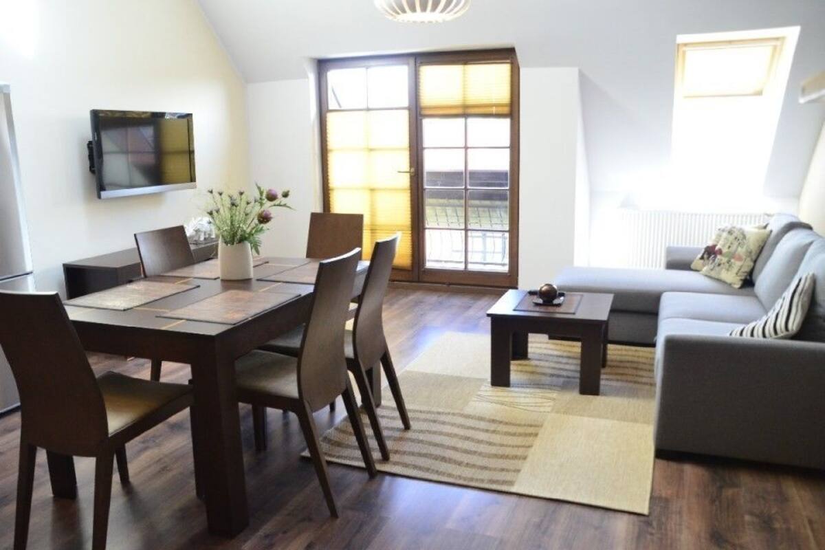 A modern living room with a dining table, chairs, a sofa, a TV mounted on the wall, and a large window with blinds.