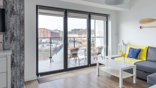 Un salón moderno con un ventanal grande, una mesa de centro blanca, un sofá gris y vistas a un canal y edificios.