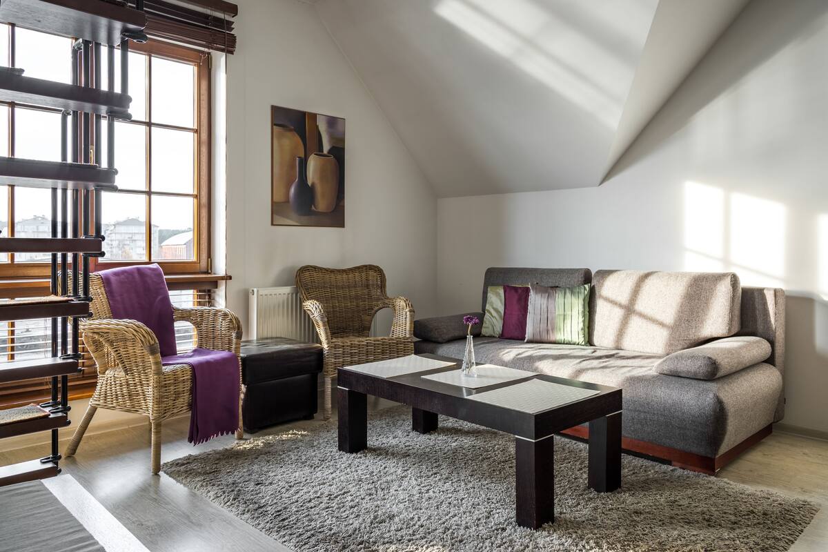 A modern living room with a high ceiling, a large window, a sofa, a coffee table, and wicker chairs.