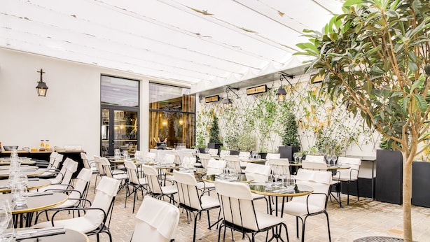 A restaurant with outdoor seating, white chairs, and tables, surrounded by plants and a tree. - Mom’Art Hotel (Paris)