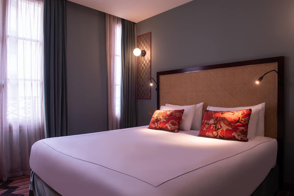 Une chambre d’hôtel avec un grand lit, deux oreillers, une tête de lit ornée d’un motif tissé, une lampe fixée au mur et une fenêtre avec des rideaux légers.