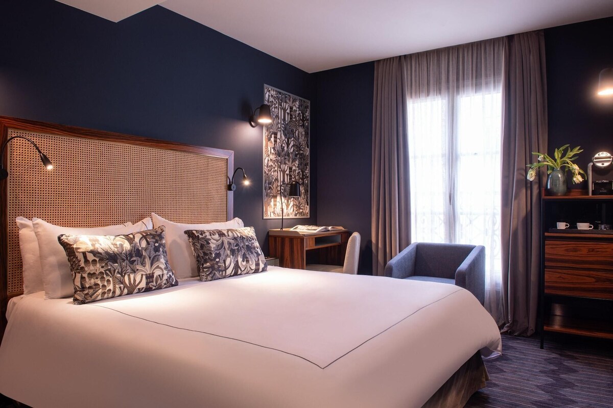 A hotel room with a large bed, a desk, a chair, a wall-mounted lamp, and a window with curtains.