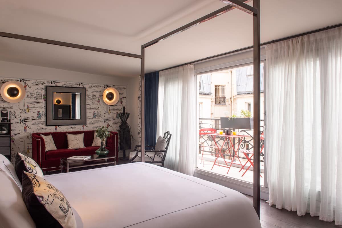 Une chambre avec un grand lit, un canapé rouge, une petite table et une chaise. Il y a un balcon avec des chaises rouges et une table à l’extérieur de la fenêtre.