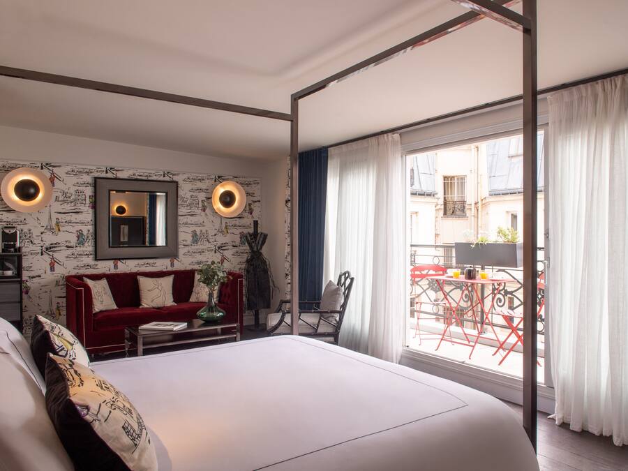 Un dormitorio con una cama grande, un sofá rojo, una mesita y una silla. Hay un balcón con sillas y una mesa rojas fuera de la ventana.