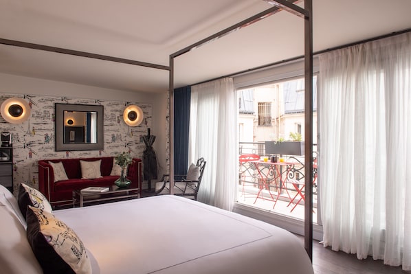 Un dormitorio con una cama grande, un sofá rojo, una mesita y una silla. Hay un balcón con sillas y una mesa rojas fuera de la ventana.
