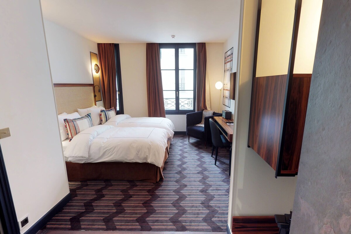A hotel room with two beds, a desk, a chair, a window with curtains, and a patterned carpet.