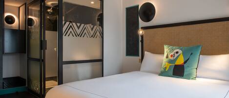 A modern hotel room with a large bed, a glass-enclosed walk-in closet, a decorative pillow, and a wall-mounted light fixture.