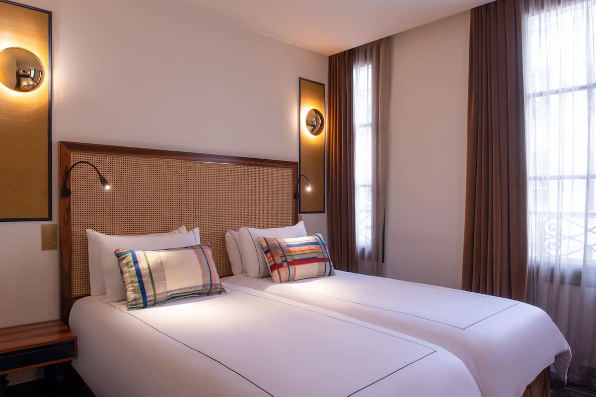 Une chambre d’hôtel avec un lit double, deux tables de chevet, une tête de lit ornée d’un motif tissé, des lampes fixées au mur et une fenêtre avec des rideaux.