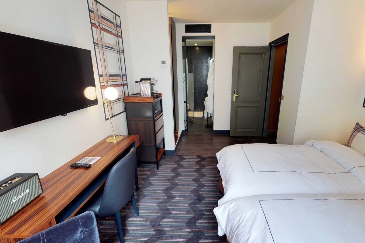 A hotel room with a bed, a desk with a chair, a TV, and a bathroom visible through an open door.