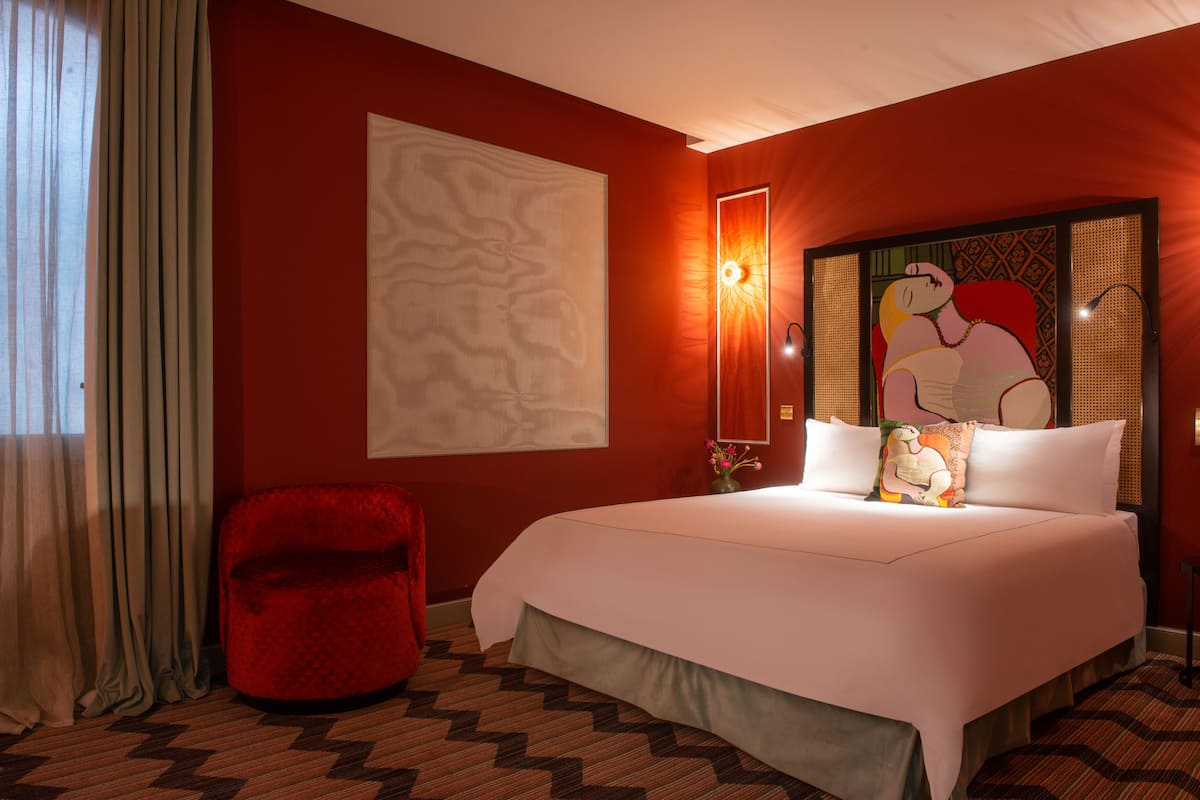 Une chambre d’hôtel avec un grand lit, un fauteuil rouge, une œuvre d’art murale et un sol à motifs.