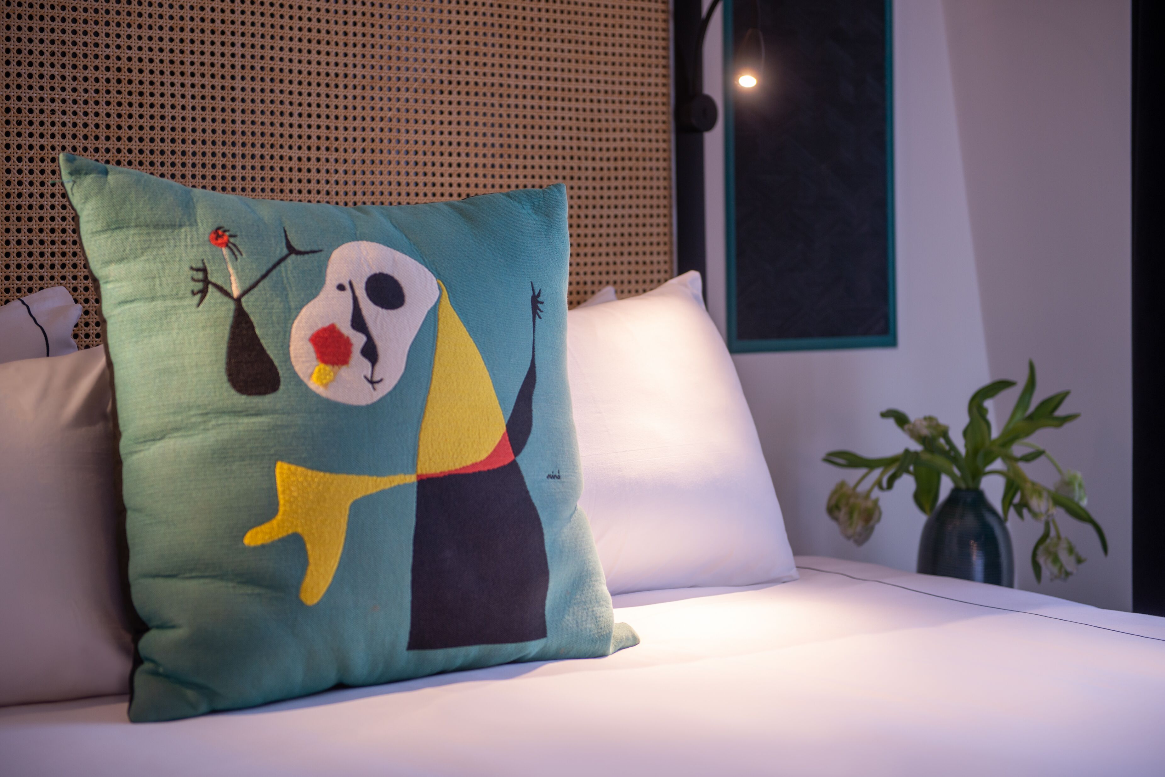 Una cama con un cojín de colores que presenta arte abstracto, un cojín blanco y un florero con flores marchitas sobre una mesa.