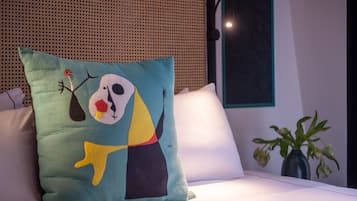 Una cama con un cojín de colores que presenta arte abstracto, un cojín blanco y un florero con flores marchitas sobre una mesa.