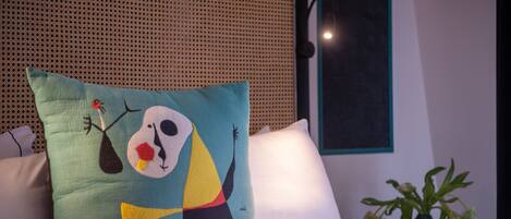 A bed with a colorful pillow featuring abstract art, a white pillow, and a vase with wilted flowers on a table.