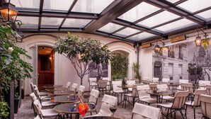 A covered outdoor café with tables and chairs, a glass ceiling, and a mural of a building. - Mom’Art Hotel (Paris)