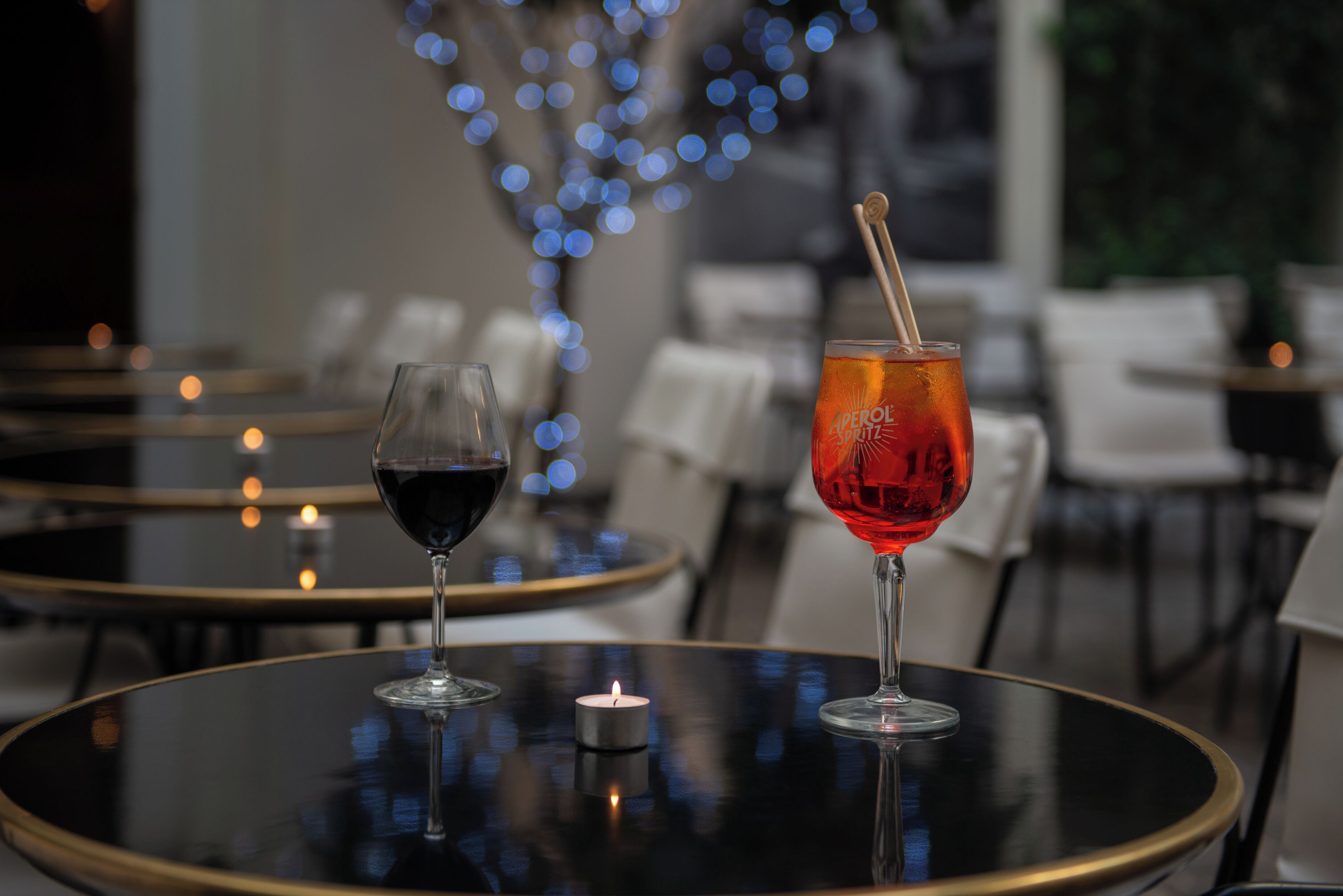 Two glasses of red wine on a round table with a lit candle, in a dimly lit restaurant with blurred background chairs and bokeh lights.