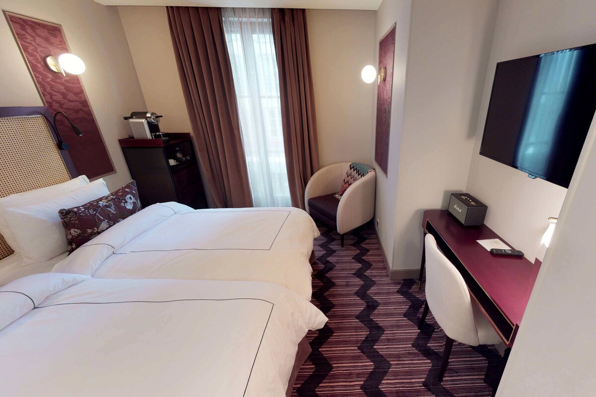 A hotel room with two beds, a desk with a TV, a chair, and a window with curtains.