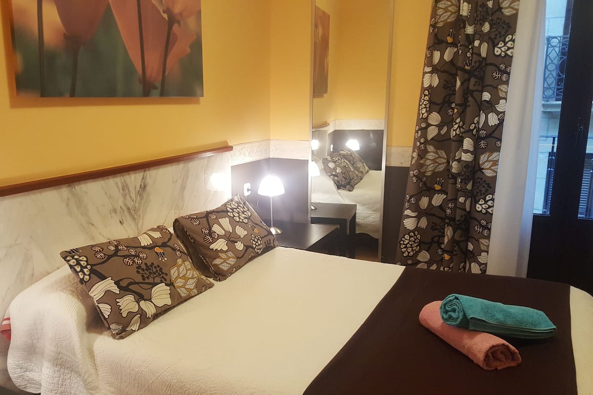 A hotel room with a bed, a nightstand, a lamp, a window with curtains, and a painting on the wall.
