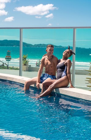 A couple sitting by a pool with a view of the ocean and mountains. - Paradiso Pero Praia Hotel (Cabo Frio)