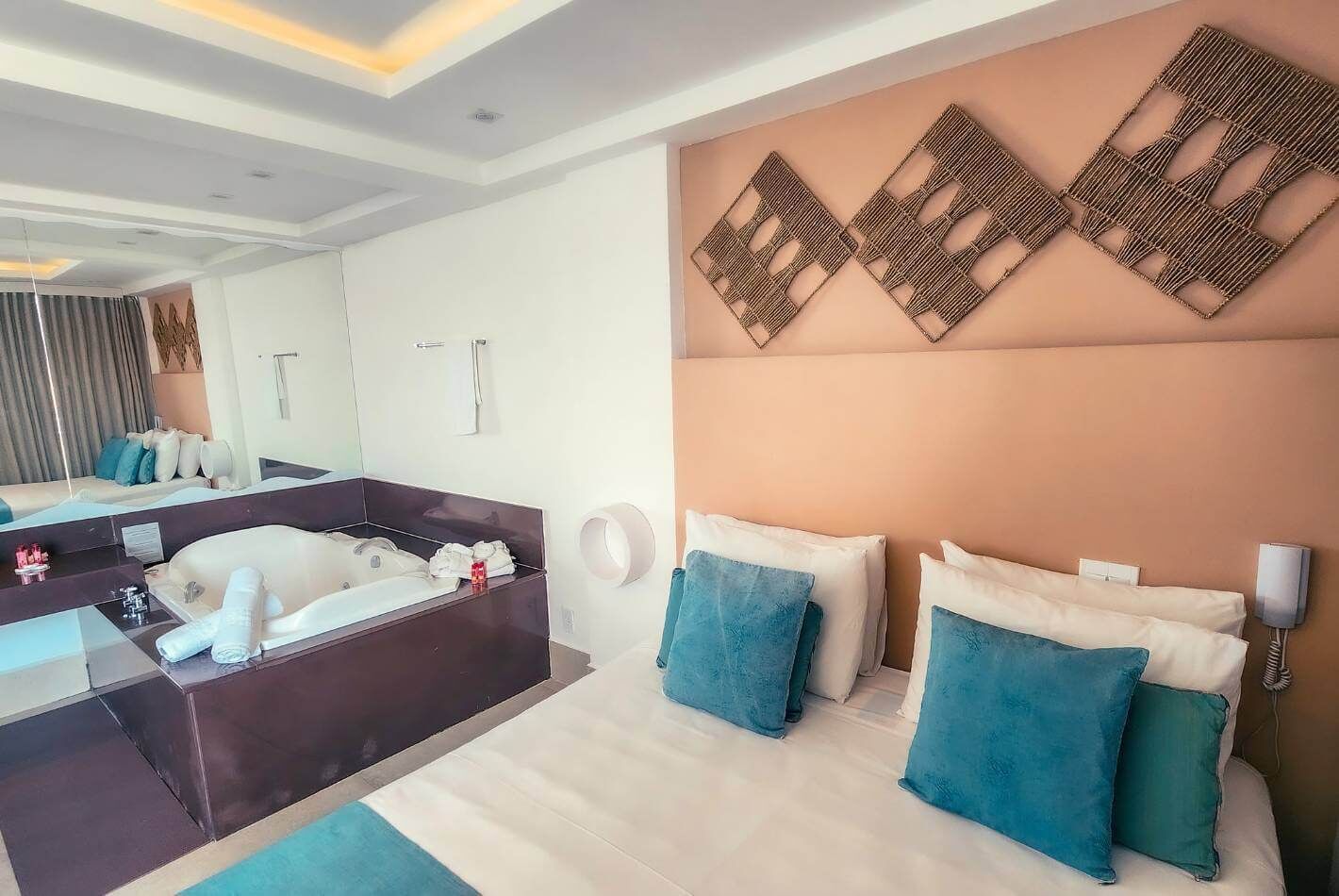 Quarto de hotel moderno com uma cama grande, banheiro separado, uma pequena escrivaninha e um detalhe decorativo na parede.