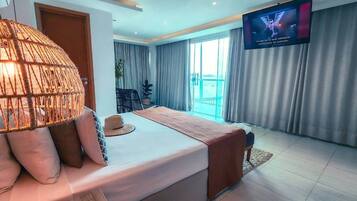 A modern hotel room with a large bed, a TV mounted on the wall, and a view of the sea through the window.