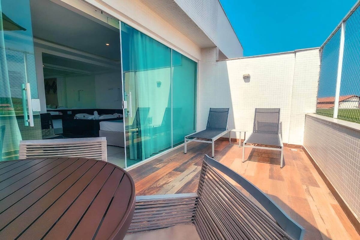 Une terrasse extérieure moderne avec une table en bois et deux fauteuils, une porte vitrée donnant sur une chambre, et un mur recouvert de carreaux blancs.