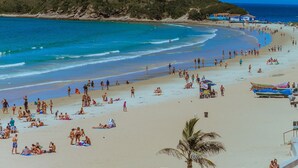 A beach with people sunbathing and swimming, a clear blue sky, and hills in the background. - Paradiso Pero Praia Hotel (Cabo Frio)