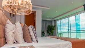 A modern hotel room with a large bed, a wooden bedside table, and a view of the sea through large windows. - Paradiso Pero Praia Hotel (Cabo Frio)