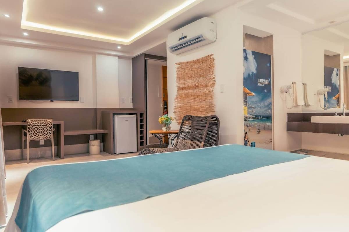 Une chambre d’hôtel moderne dotée d’un grand lit, d’un bureau avec une chaise, d’un petit réfrigérateur, d’une télévision et d’une salle de bain avec un miroir et un lavabo.