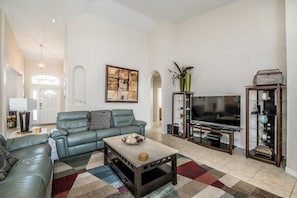A spacious living room with a large sofa, a coffee table, a flat-screen TV, and a bookshelf.