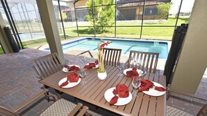 An outdoor dining area with a pool view, wooden table and chairs, and a vase with flowers. - Providence 2323EVDIL (Davenport)
