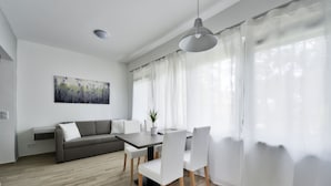 A modern living room with a dining area, featuring a grey sofa, white chairs, a dining table, and a large window with sheer curtains. - Villa Oliver 2 Siófok (Siófok)